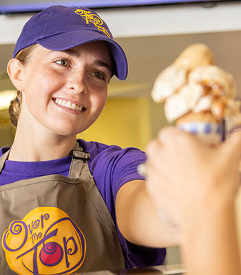 A smiling Over The Top Ice Cream scooper handing a cone to a customer over the counter.