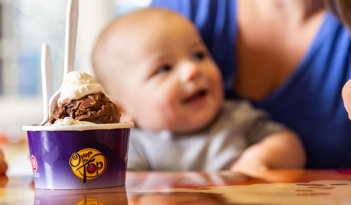 A double scoop of Over The Top Ice Cream sits on a table in front of an out-of-focus baby.