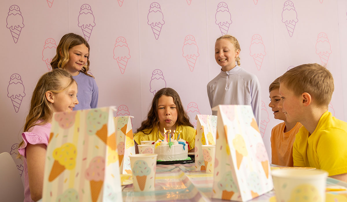 A birthday girl blows out the candles on her ice cream cake at the Over The Top Ice Cream party room, surrounded by friends.