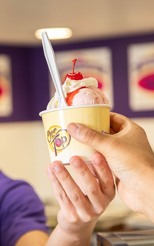 An Over The Top Ice Cream scooper hands a customer a scoop of ice cream in a branded paper cup over the counter.